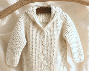 May include: Cream-colored, hooded baby cardigan with long sleeves and a button-up front. The knitted sweater is displayed on a wooden hanger, showcasing its soft texture and design. The neutral color makes it suitable for various outfits.