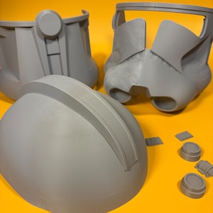 May include: Gray 3D-printed helmet parts, including a helmet dome, face mask, and other components, arranged on a yellow surface. The pieces are detailed and appear to be from a science fiction or cosplay project.