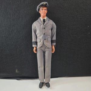 May include: A vintage doll dressed in a gray uniform, including a jacket, pants, and a cap. The doll has a tie and black shoes. The uniform has black stripes on the sleeves. The doll is standing against a dark background.