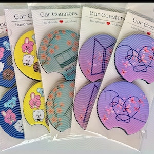 Puede incluir: Una colección de posavasos para coche con varios diseños. Los posavasos presentan personajes de dibujos animados, estampados florales y formas geométricas en tonos azules, amarillos y morados. Cada posavasos está empaquetado en una funda de plástico transparente.