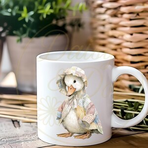 May include: White ceramic mug with a duckling illustration wearing a bonnet and patchwork coat. The duckling has a yellow beak and is standing. The mug has a curved handle. The background is blurred, with plants and a wicker basket.