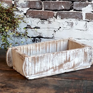 Könnte beinhalten: Ein weiß gestrichener Holz-Pflanztopf mit rustikalem, abgenutztem Finish. Die Box hat eine rechteckige Form mit abgerundeten Kanten und einem leicht erhöhten Rand oben.
