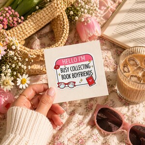 May include: A rectangular white sticker featuring a pink header with the text HELLO I'M in white, followed by the black text BUSY COLLECTING BOOK BOYFRIENDS. The sticker includes illustrations of red lips, red-rimmed glasses, and a red book with a heart.