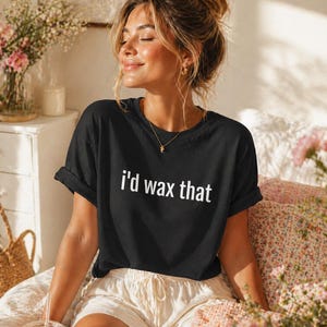 May include: Black t-shirt with the text "i'd wax that" in white. The shirt is worn with white shorts. The image shows a person sitting on a bed with flowers and a drink.