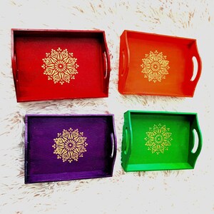 Puede incluir: Cuatro bandejas de servir de madera con dise&ntilde;os pintados en oro. Las bandejas son rojas, naranjas, moradas y verdes.