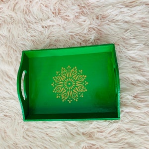 Puede incluir: Una bandeja de servir de madera verde con un dise&ntilde;o de mandala dorado. La bandeja tiene un borde elevado y dos asas.