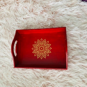 Puede incluir: Una bandeja de servir de madera roja con un dise&ntilde;o de mandala dorado. La bandeja tiene un asa a cada lado.