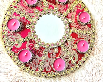 Puede incluir: Una bandeja redonda decorativa con un espejo en el centro. La bandeja está decorada con detalles dorados y rosados y tiene siete pequeños portavelas alrededor del espejo.