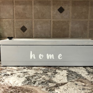 May include: A light gray wooden box with a clear lid. The box has the word "home" written on the front in white lettering.
