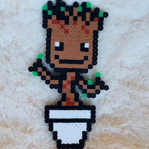May include: A brown and green pixel art character of Groot from Guardians of the Galaxy, standing in a white pot. The character has a small smile and is holding out its arms.