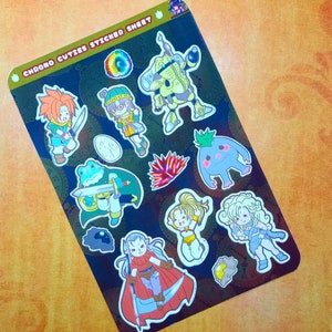 May include: A sticker sheet with the text "CHRONO CUTIES STICKER SHEET" features colorful cartoon characters. The stickers include a frog with a sword, a robot, and other fantasy figures. The background is dark with a clock pattern.