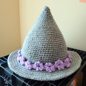 K&ouml;nnte beinhalten: Ein grauer H&auml;kelhut mit einem violetten Blumenbesatz. Der Hut liegt auf einer schwarzen Oberfl&auml;che.