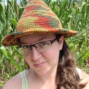 K&ouml;nnte beinhalten: Eine Person tr&auml;gt einen gr&uuml;nen, orangefarbenen und gelben H&auml;kelhut mit breiter Krempe. Der Hut befindet sich in einem Feld mit hohen gr&uuml;nen Maispflanzen.