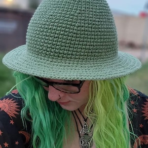 K&ouml;nnte beinhalten: Eine Person mit gr&uuml;nen und gelben Haaren tr&auml;gt einen gr&uuml;nen H&auml;kelhut. Sie tr&auml;gt ein schwarzes Shirt mit einem Sonnen- und Mondmotiv und eine silberne Halskette mit einem Mond- und einem Triquetra-Anh&auml;nger.