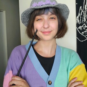 K&ouml;nnte beinhalten: Eine Person tr&auml;gt einen grauen und lila gestrickten Hexenhut mit einem schwarzen Zauberstab in der Hand. Sie tr&auml;gt einen mehrfarbigen Cardigan mit rosa, lila, blauen, gr&uuml;nen und gelben Streifen.
