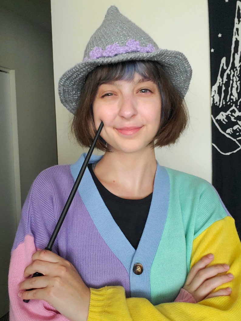 K&ouml;nnte beinhalten: Eine Person tr&auml;gt einen grauen und lila gestrickten Hexenhut mit einem schwarzen Zauberstab in der Hand. Sie tr&auml;gt einen mehrfarbigen Cardigan mit rosa, lila, blauen, gr&uuml;nen und gelben Streifen.