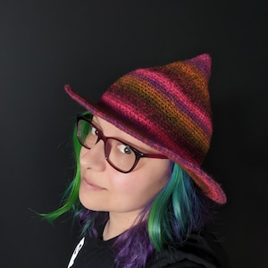 K&ouml;nnte beinhalten: Eine Person tr&auml;gt einen rot-orange-violetten gestreiften Strick-Hexenhut mit breiter Krempe. Sie hat gr&uuml;n-violettes Haar und tr&auml;gt eine Brille.