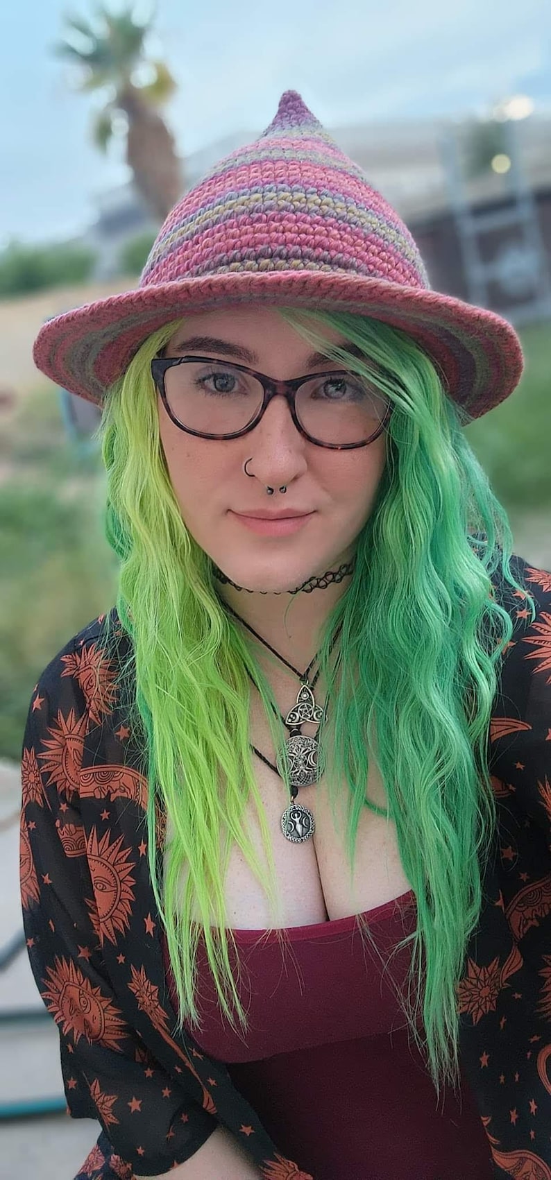 K&ouml;nnte beinhalten: Eine Person mit gr&uuml;nen Haaren tr&auml;gt einen rosa-violetten gestreiften H&auml;kelhut, eine schwarze Brille, eine schwarze Halskette und mehrere silberne Halsketten mit Anh&auml;ngern. Sie tr&auml;gt ein schwarz-rotes gemustertes Hemd mit tiefem Ausschnitt.