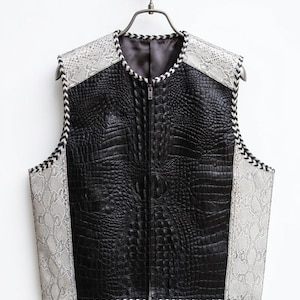 May include: A black leather vest with a crocodile texture on the front and a snakeskin pattern on the shoulders and sides. The vest has a zipper closure and is trimmed with a black and white zigzag pattern.