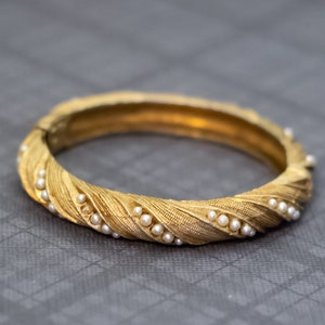 May include: A gold-colored hinged bangle bracelet with a textured, twisted design. Small, white pearls are set along the twisted pattern. The bracelet is shown on a gray surface.