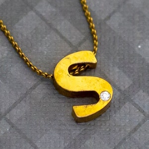 Puede incluir: Un collar de cadena de oro con un colgante de oro con la letra 'S' con un pequeño diamante.