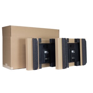 May include: Two black flat-screen TVs are packaged in cardboard boxes with protective foam inserts. A large, unopened cardboard box is on the left. The TVs are facing away, revealing their ports and mounting hardware.