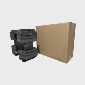 May include: A rectangular brown cardboard shipping box with the text theBOX larger printed in black and orange on the front. Beside the box is a black electronic device secured between two thick, dark gray foam protective packaging blocks.