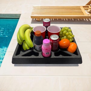 May include: A black tray holds a bunch of bananas, green grapes, two oranges, and various beverages, including a pink drink, a dark juice bottle, and two cans of soda. Two red cups are also in the tray. The tray is on a white tiled surface near a pool.