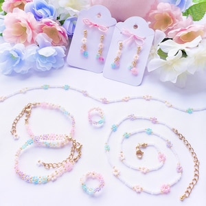 May include: A set of pastel-colored flower bead jewelry, including a necklace, bracelet, and rings. The jewelry is displayed on a white surface with a pink plush bunny and flowers.