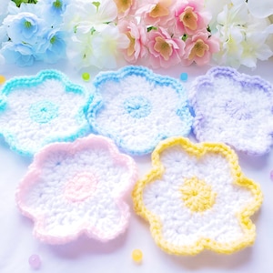 May include: Five crocheted flower-shaped coasters in pastel colors. The coasters are white with a light blue, light purple, light pink, and yellow center.