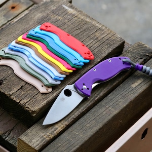 May include: A purple folding knife with a silver blade and a lanyard, next to a stack of colorful knife scales in red, blue, green, yellow, and other colors. The items are arranged on weathered wooden planks.