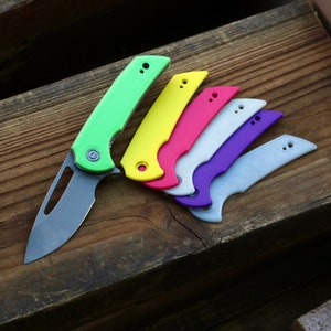 May include: A set of six colorful knife scales, including green, yellow, pink, white, purple, and gray. The scales are arranged in a fan shape on a wooden surface.