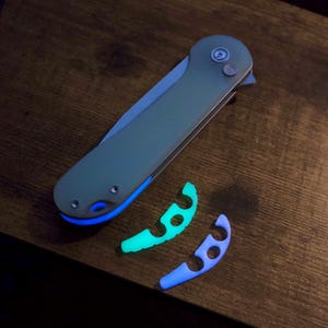 May include: A silver pocket knife with a blue glow-in-the-dark handle. The knife is lying on a wooden surface with two glow-in-the-dark blue and green plastic pieces.