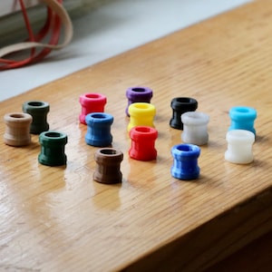 May include: Assortment of colorful plastic spools, arranged in a grid pattern on a wooden surface. The spools are various colors, including red, blue, green, yellow, white, black, brown, and purple.