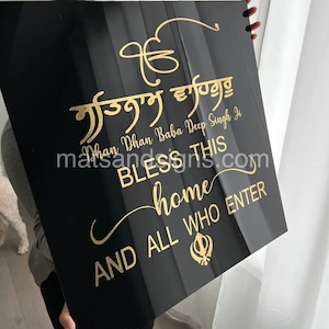 May include: A black rectangular sign with gold lettering. The sign features text in a stylized script, including the words "BLESS THIS home AND ALL WHO ENTER." The sign is held up by a person.