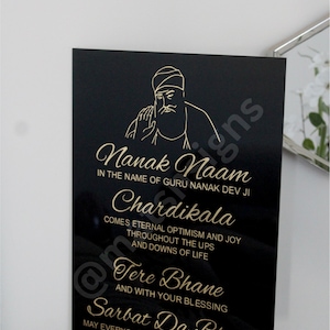 Puede incluir: Placa rectangular negra con letras doradas e ilustración de una figura religiosa. El texto incluye frases como "Nanak Naam" y "Chardikala". La placa se muestra en una pared blanca.