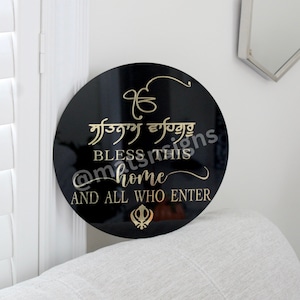 Puede incluir: Un letrero redondo negro con letras doradas que dice "BLESS THIS HOME AND ALL WHO ENTER". El letrero presenta símbolos religiosos sijes y caligrafía. El letrero está en una pared blanca.