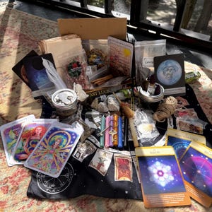 May include: A collection of spiritual items including tarot cards with colorful illustrations, small glass jars with metal lids, assorted colored taper candles, a miniature broom, dried herbs in plastic bags, a metal key, and a cardboard box labeled Fast Luck.