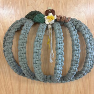 May include: A handmade macrame pumpkin wreath in shades of gray-green. The wreath features three connected loops, with a crocheted stem, leaves, and a flower in shades of green, brown, and yellow. The wreath is designed for seasonal home decor.