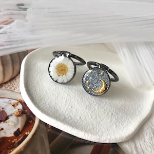 Puede incluir: Dos soportes de anillo para teléfono con un anillo de metal negro y una parte superior de resina transparente. Un soporte presenta una margarita blanca con un centro amarillo. El otro soporte presenta un fondo de purpurina plateada con una luna creciente dorada y estrellas.
