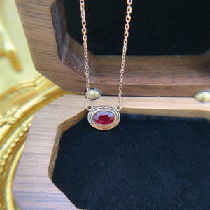 May include: A delicate rose gold necklace featuring a single oval-cut deep red ruby gemstone set in a smooth, polished metal bezel. The pendant is suspended from a fine, matching rose gold chain, creating a refined and minimalist jewelry piece.