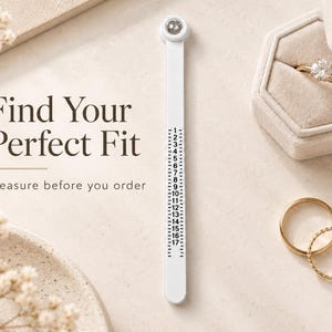 May include: A white plastic ring sizer tool with a numbered scale from 1 to 17, positioned vertically next to a beige velvet ring box containing a gold ring with a clear stone, and two additional gold rings resting nearby on a flat surface.