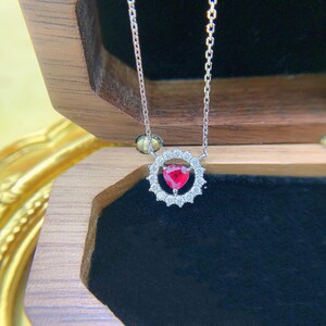 May include: A delicate silver-toned chain necklace featuring a pendant with a central heart-shaped red gemstone surrounded by a halo of small, clear, round-cut stones arranged in a circular, scalloped-edge setting.