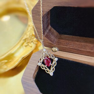 May include: A gold necklace featuring a diamond-shaped pendant with an oval-cut red ruby at the center, surrounded by intricate gold filigree metalwork and four small round-cut white diamonds set at the corners of the frame.
