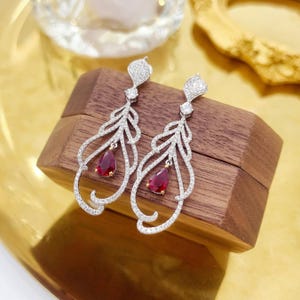 May include: A pair of silver-toned dangle earrings featuring a delicate, openwork feather-like design encrusted with small clear stones. Each earring is centered with a single, teardrop-shaped deep red stone set in a gold-toned metal base.