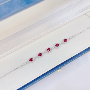 May include: A silver-toned bracelet featuring five oval-cut deep red rubies, each surrounded by a halo of small clear stones. The chain consists of delicate silver links interspersed with additional small clear stones, creating a refined and symmetrical design.