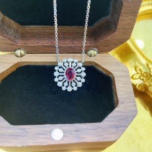 May include: A silver-toned necklace featuring a central oval-cut deep red gemstone surrounded by a sunburst-style halo of small, round, clear stones. The pendant is suspended from a delicate silver-toned chain, creating a radiant, circular design.