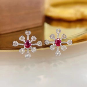 May include: A pair of silver stud earrings featuring a central oval-cut red ruby surrounded by a starburst arrangement of small, round, rose-cut white diamonds set on thin silver spokes, creating a symmetrical, multi-pointed floral design.
