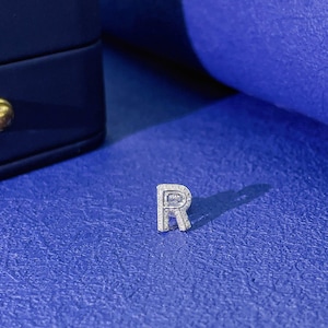 May include: A silver-toned letter R charm featuring a pavé-style surface covered in small, clear, reflective stones. The letter has a block-style font with a metallic border and a small, rectangular attachment loop at the top for hanging.