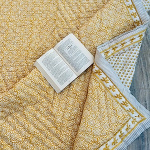 Puede incluir: Un edredón con estampado floral amarillo y blanco con un libro encima. El edredón tiene un diseño floral repetido y un borde a rayas con un patrón floral. Un libro abierto con texto es visible.